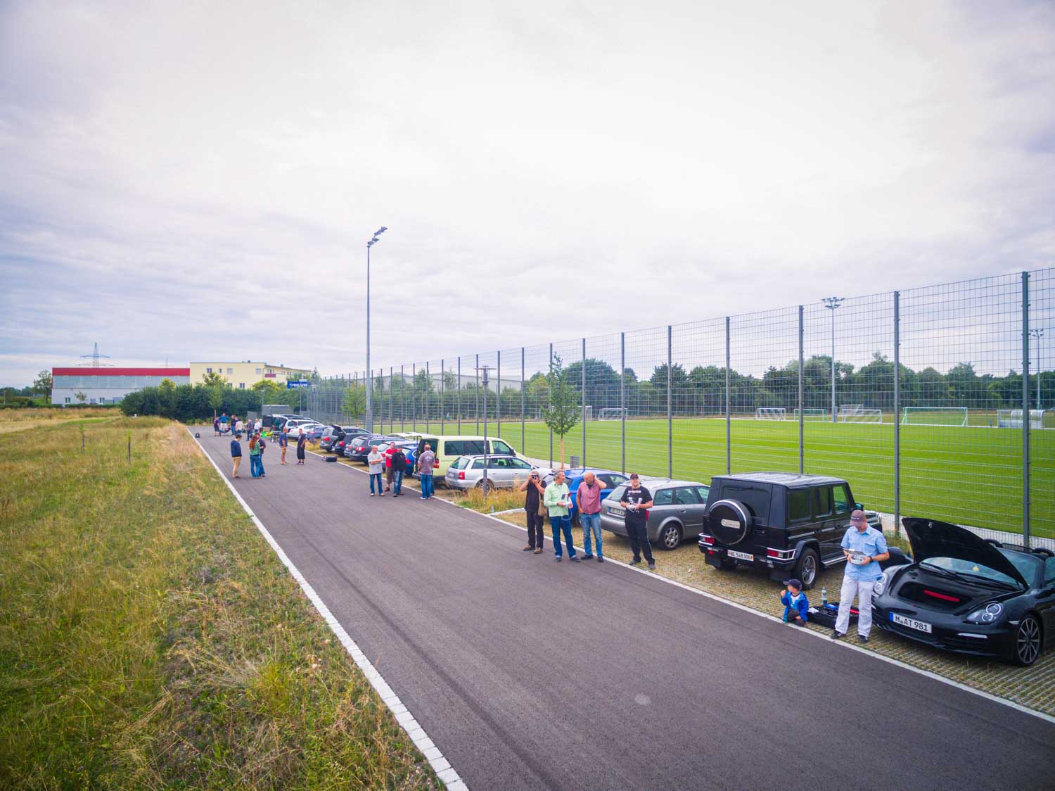 People gather near parked cars beside a fenced soccer field, with a cloudy sky in the background.