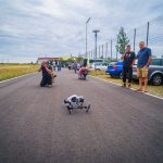 Man photographing a small drone on an outdoor road with people and parked cars in the background.