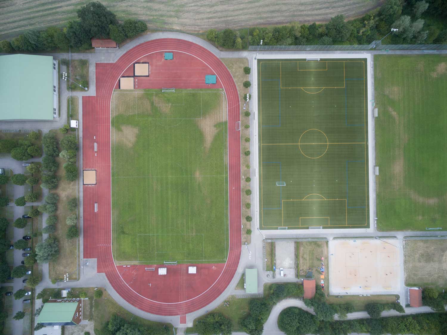 Aerial view of sports complex with red running track, green soccer field, and adjacent buildings surrounded by greenery.