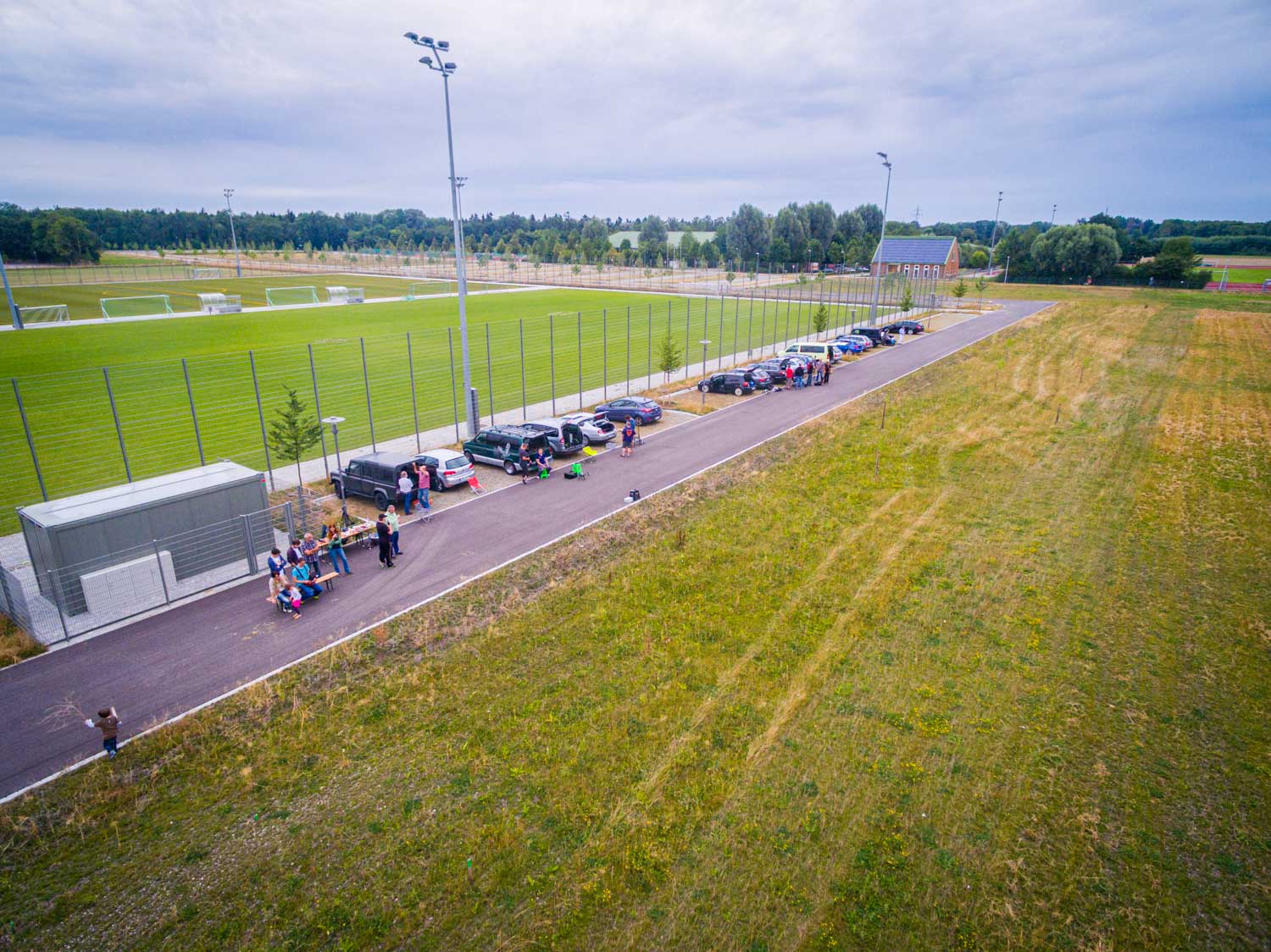 Parking area beside a sports field with people gathered and cars lined up, surrounded by green landscape and fields.