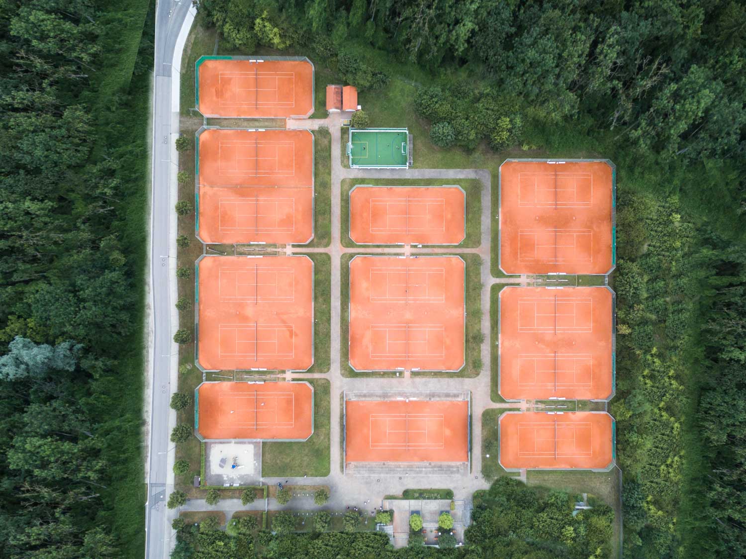 Aerial view of multiple clay tennis courts surrounded by lush green forest and a nearby road.