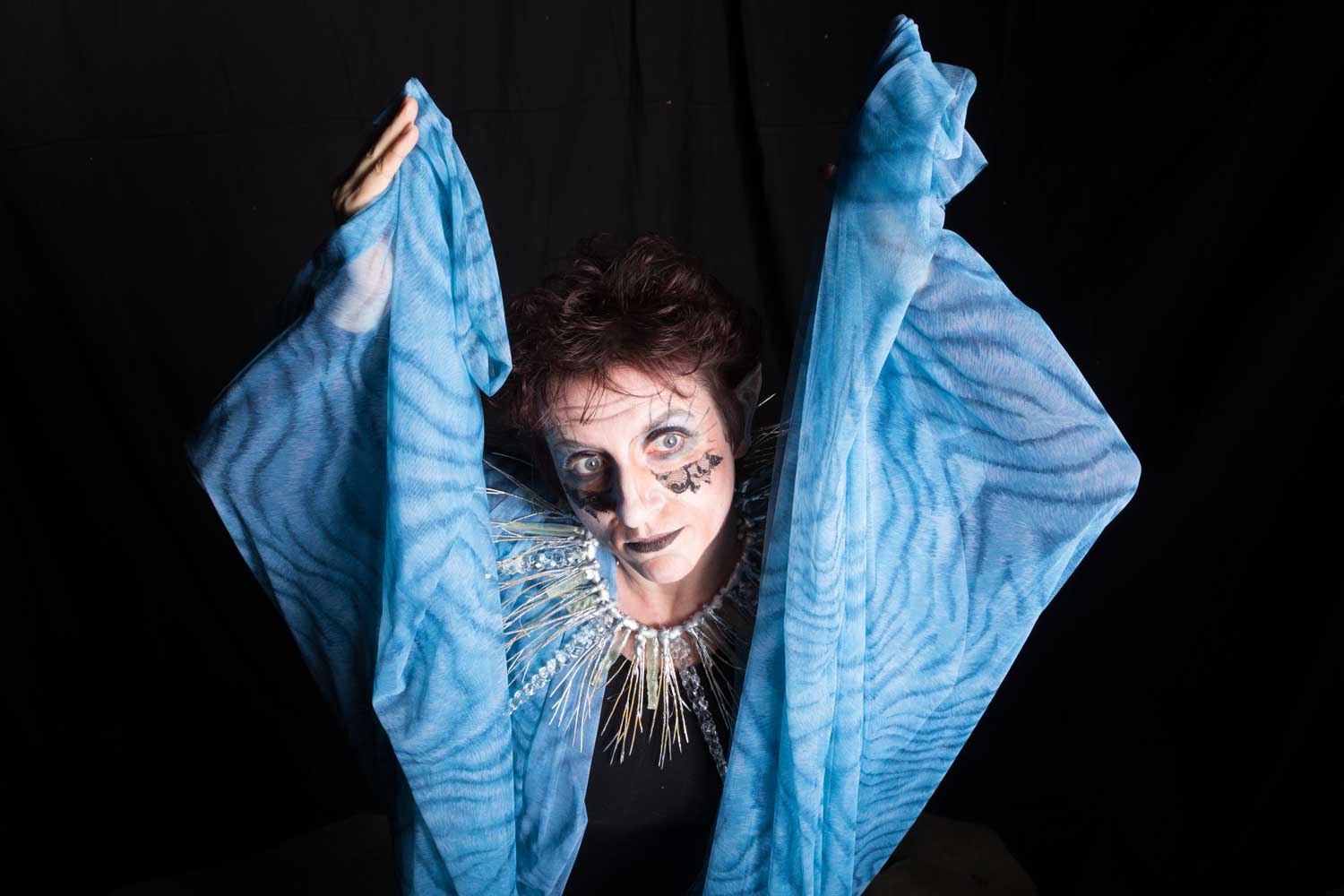 Person in blue costume with dramatic makeup and spiky collar poses against a dark background.