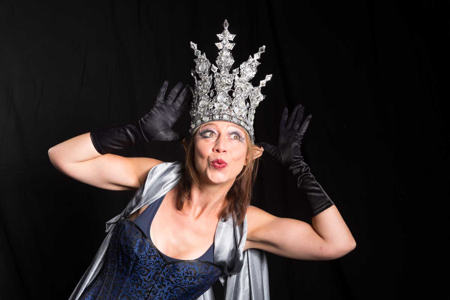 Woman in black gloves and ornate silver crown makes a playful face against dark background.