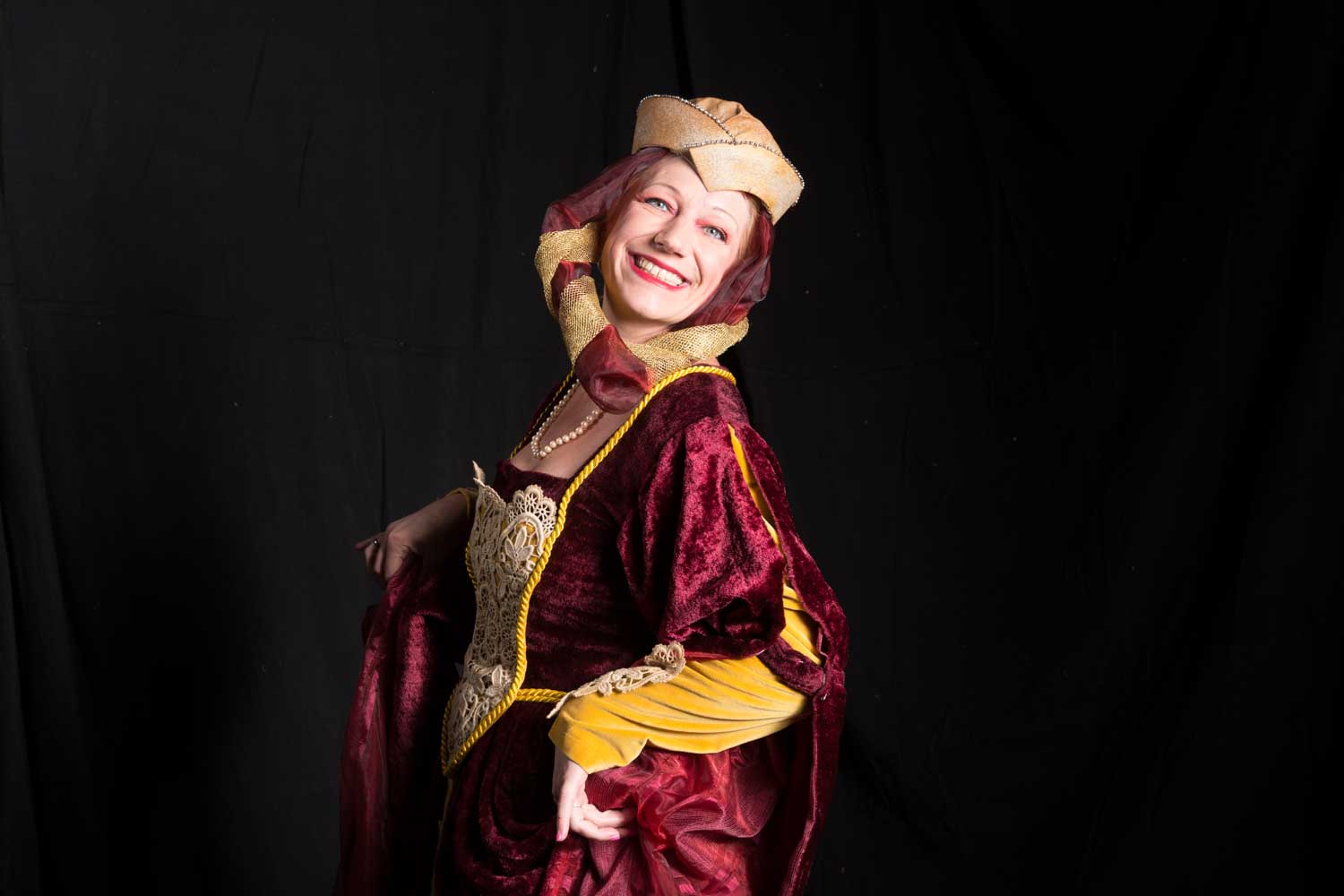 Smiling woman in a red medieval costume with a gold crown poses against a dark background.