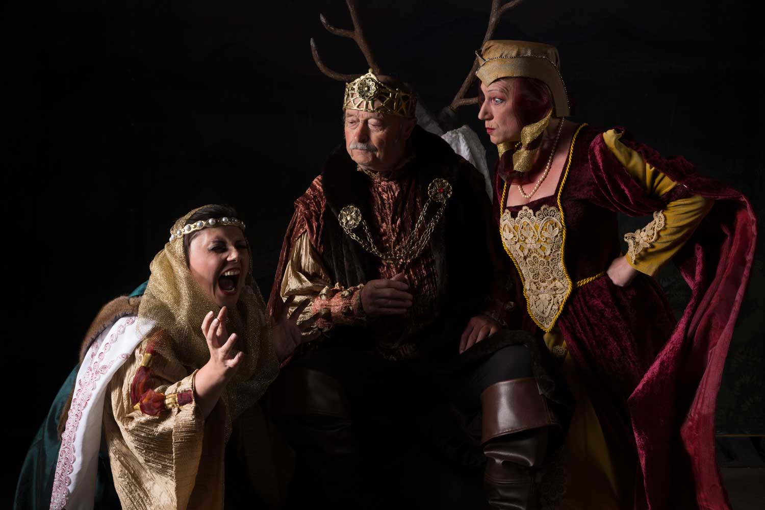 Three actors in medieval costumes perform dramatically on stage, featuring rich fabrics and regal accessories.