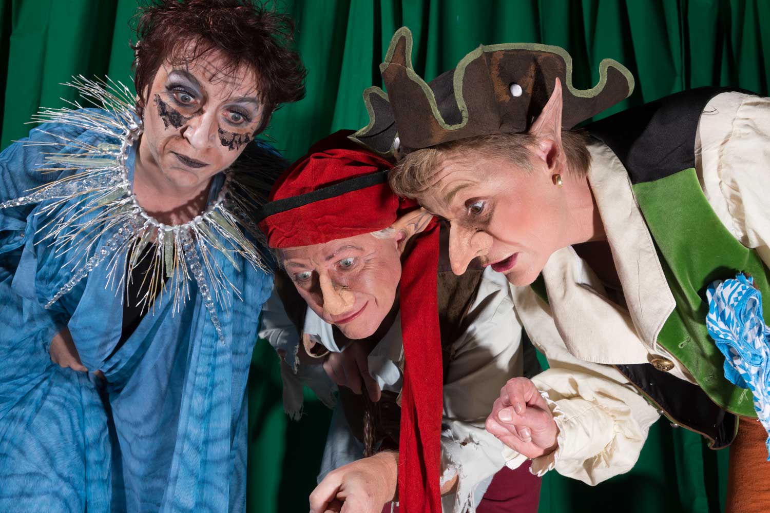 Three actors in elaborate fantasy costumes and makeup perform on stage with a green curtain backdrop.