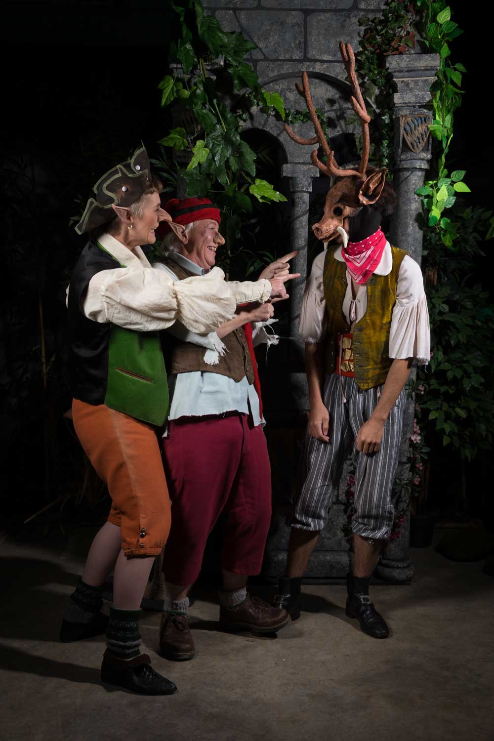 Three people in elf costumes laugh next to a person wearing a reindeer mask, standing in front of a stone archway.