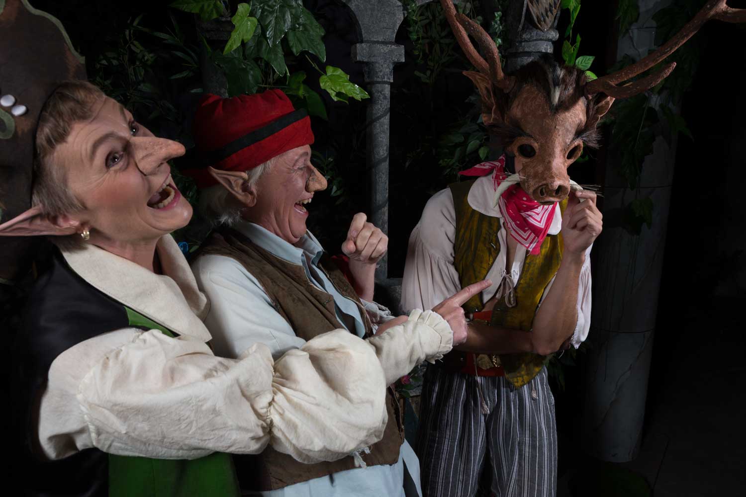 People in fantasy costumes laughing, one wearing a deer mask with antlers, in a forest-themed setting.
