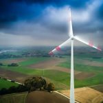 Wind turbine spinning over vast green and brown farmland under a cloudy sky.