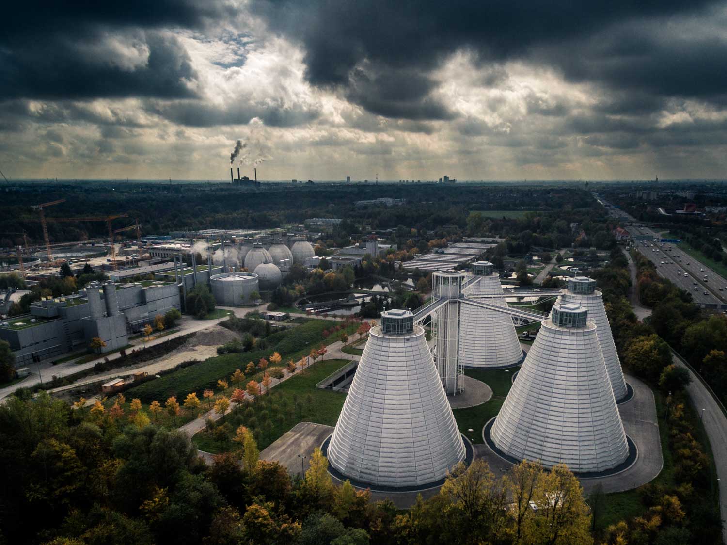 Aerial view of a wastewater treatment plant with cone-shaped buildings under a cloudy sky, surrounded by greenery and roads.