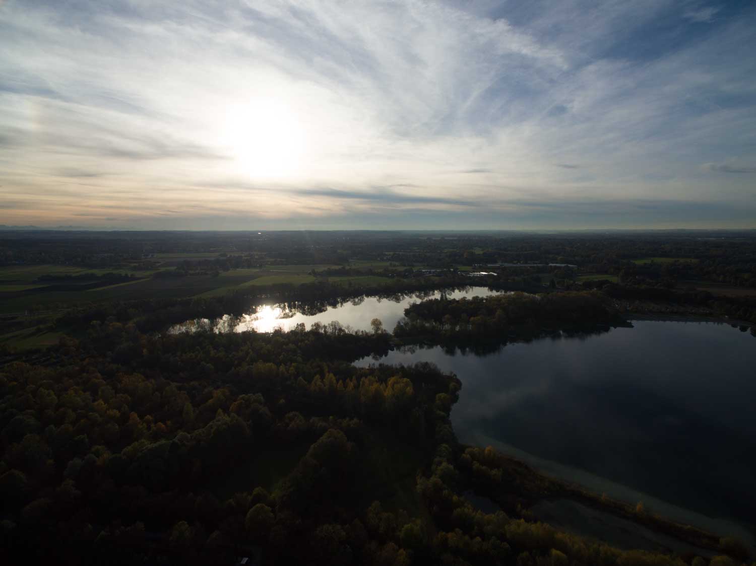 Aerial view of a serene lake reflecting sunlight, surrounded by lush green forest under a cloudy sky.