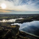Aerial view of a serene lake reflecting the cloudy sky, surrounded by forests and fields under a setting sun.
