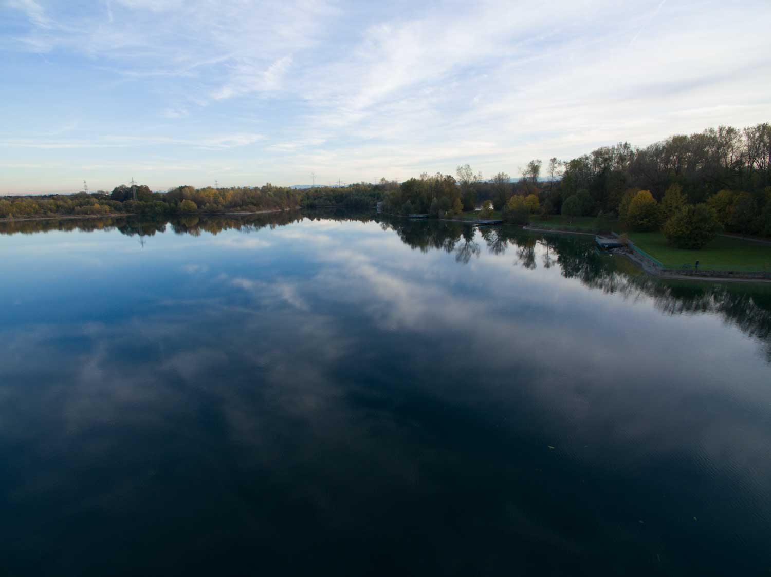 Calm lake reflecting blue sky and clouds, surrounded by autumn trees and a grassy park area.
