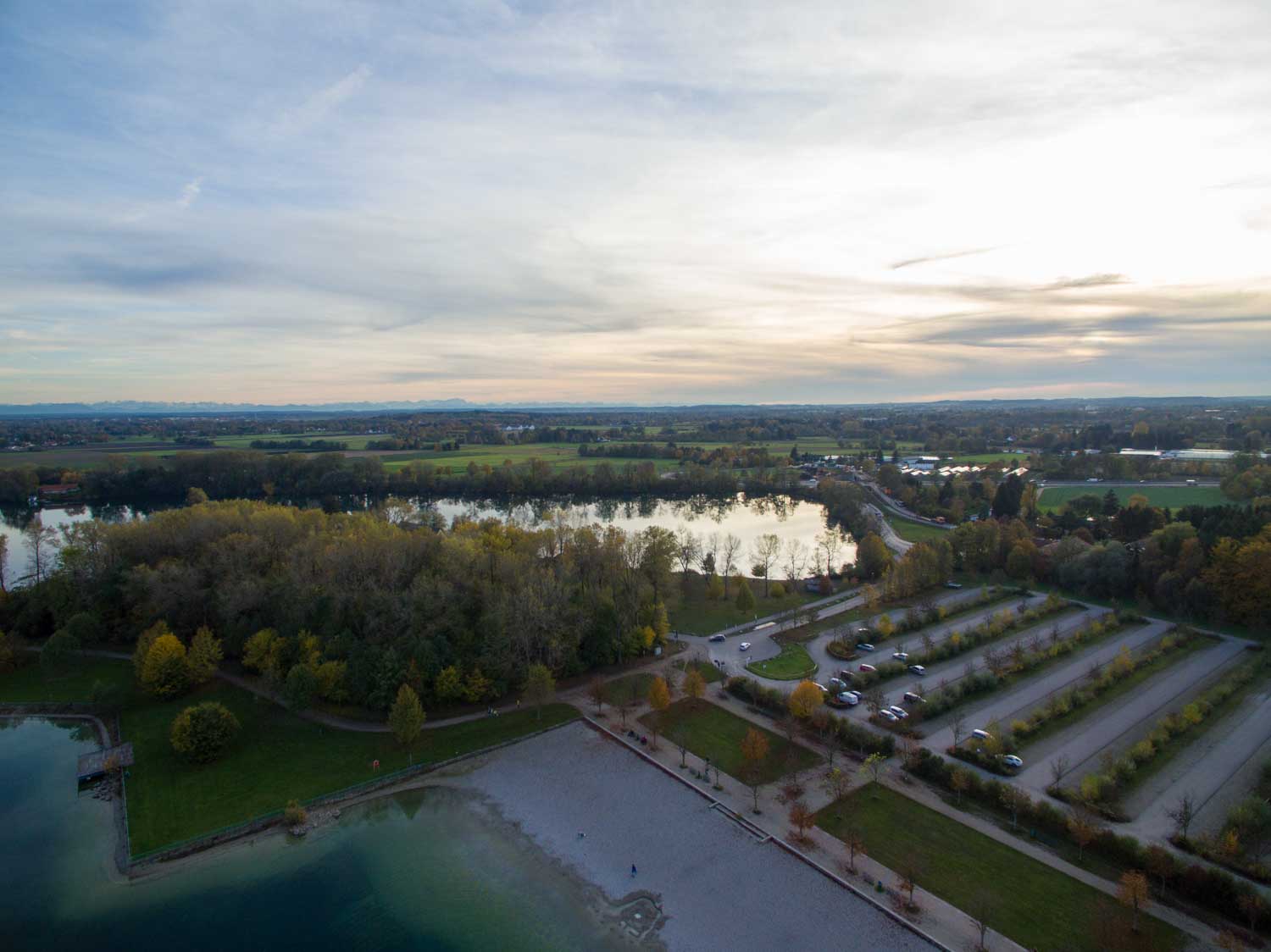 Aerial view of a serene lake surrounded by autumn trees, an empty parking lot, and a distant horizon under a cloudy sky.