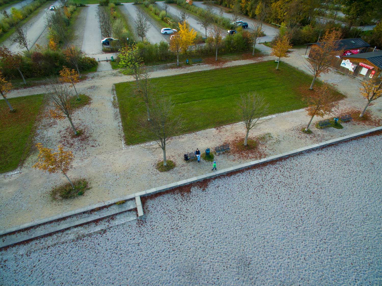 Aerial view of a park with autumn trees, grass, pathways, parking and people walking along a stone promenade.