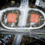 Aerial view of two adjacent red clay tennis courts surrounded by trees and roads, near a railway line.