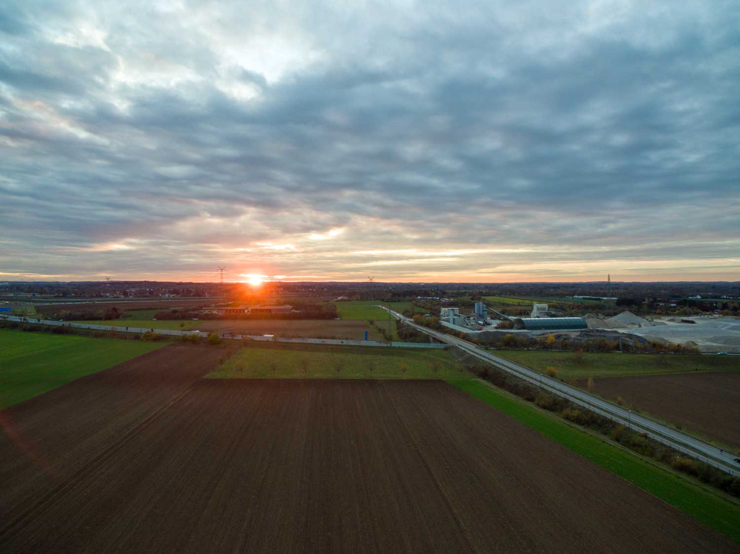 Aerial view of farmland and road at sunset with cloudy sky, wind turbines in the distance, and buildings on the horizon.