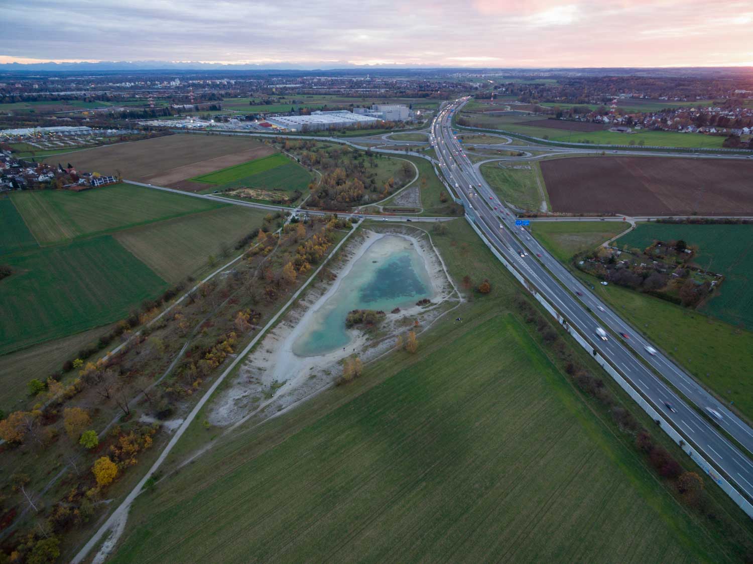Aerial view of highways intersecting near a small lake with surrounding fields and distant city skyline at sunset.