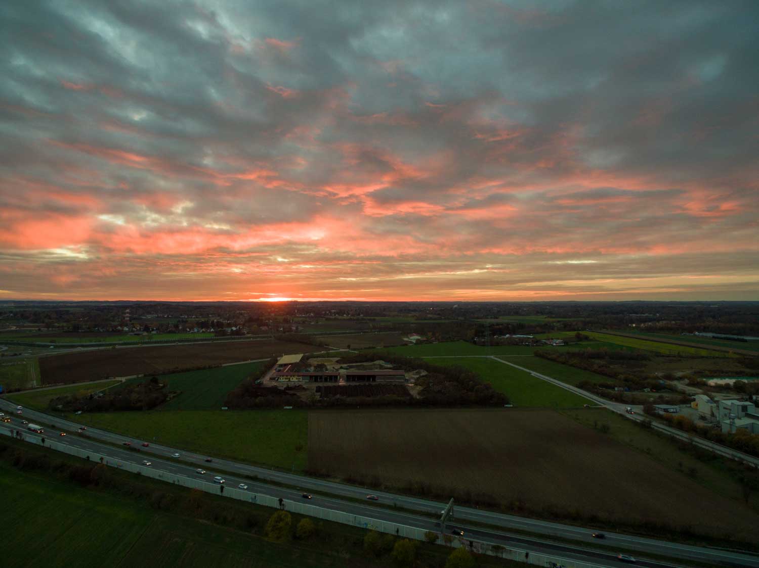 Sunset over green fields and a highway, with vibrant red and orange clouds covering the sky.