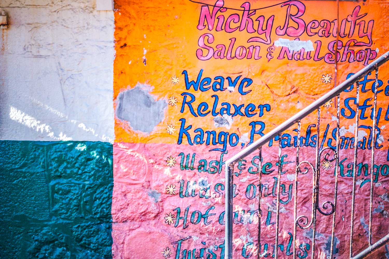 Colorful beauty salon sign on a wall, offering services like weaves, relaxers, and braids with decorative iron railing.