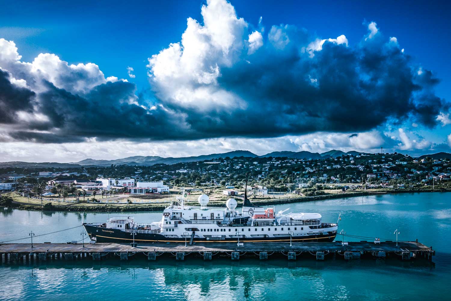 Cruise ship docked at a pier with a cloudy sky and coastal town in the background.