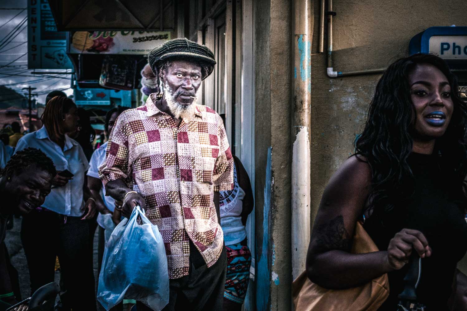 Elderly man in checkered shirt carries plastic bag on busy street, with diverse group of people in urban setting.