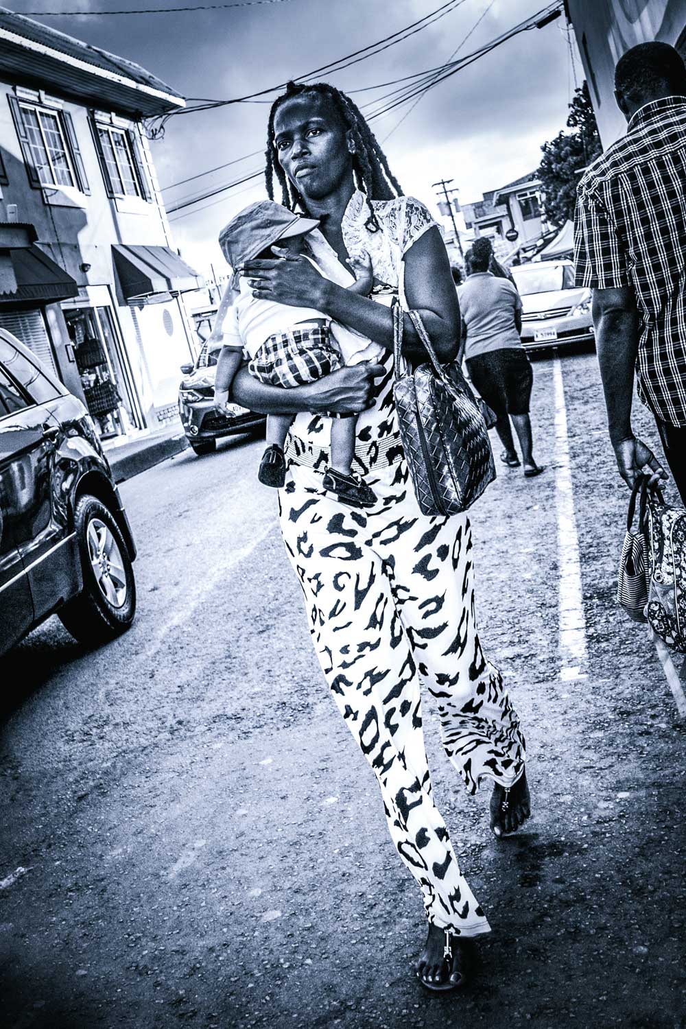 Woman carrying a baby on a street with cars and people; dressed in patterned pants, holding a bag.