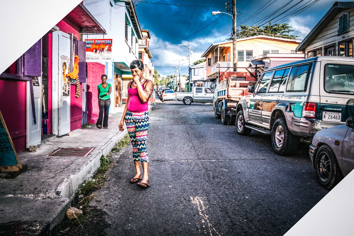 Woman in bright outfit walks on a colorful street with cars and shops on a sunny day.