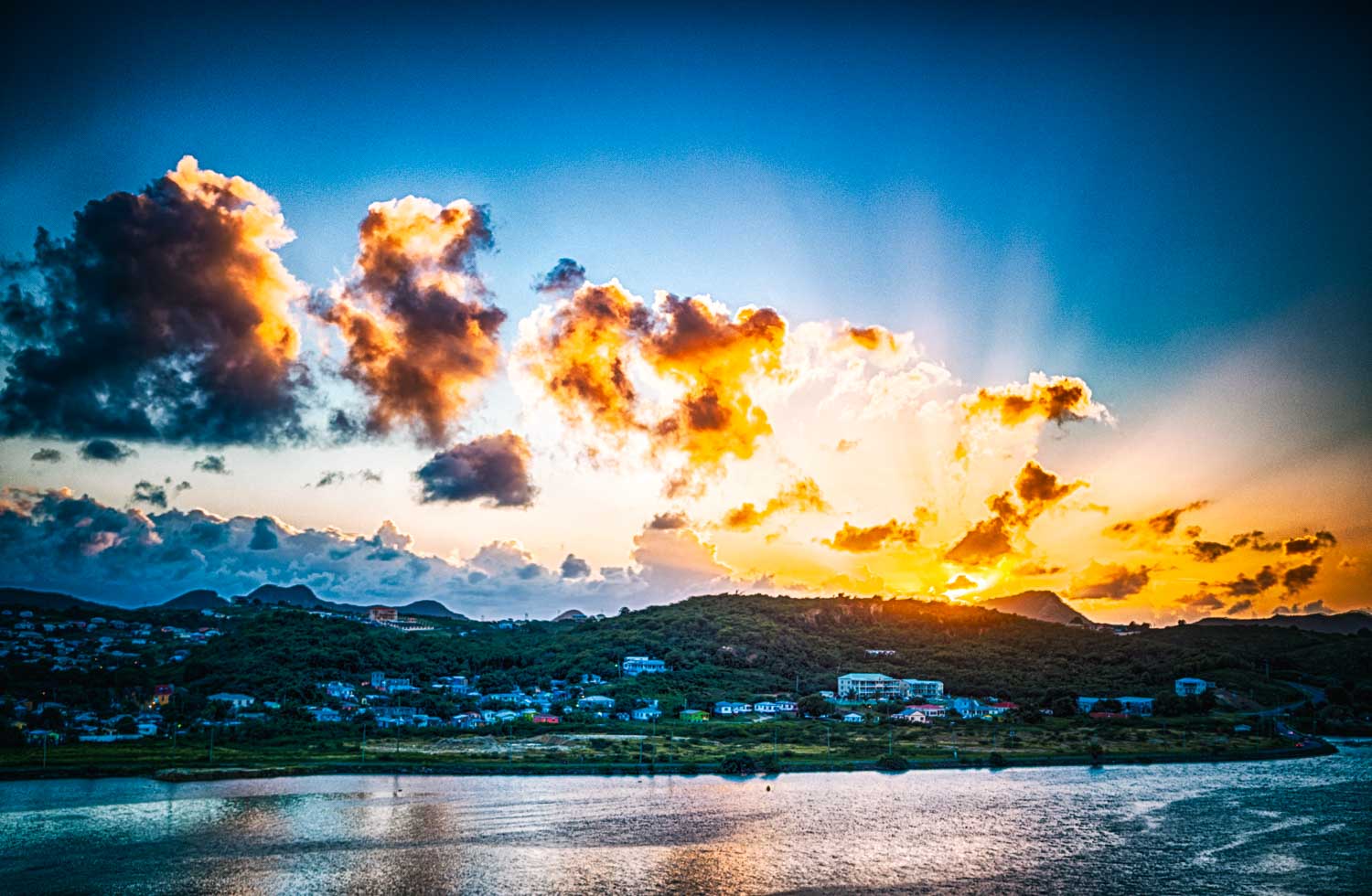 Stunning sunset over a coastal town with vibrant clouds and sun rays illuminating the horizon.