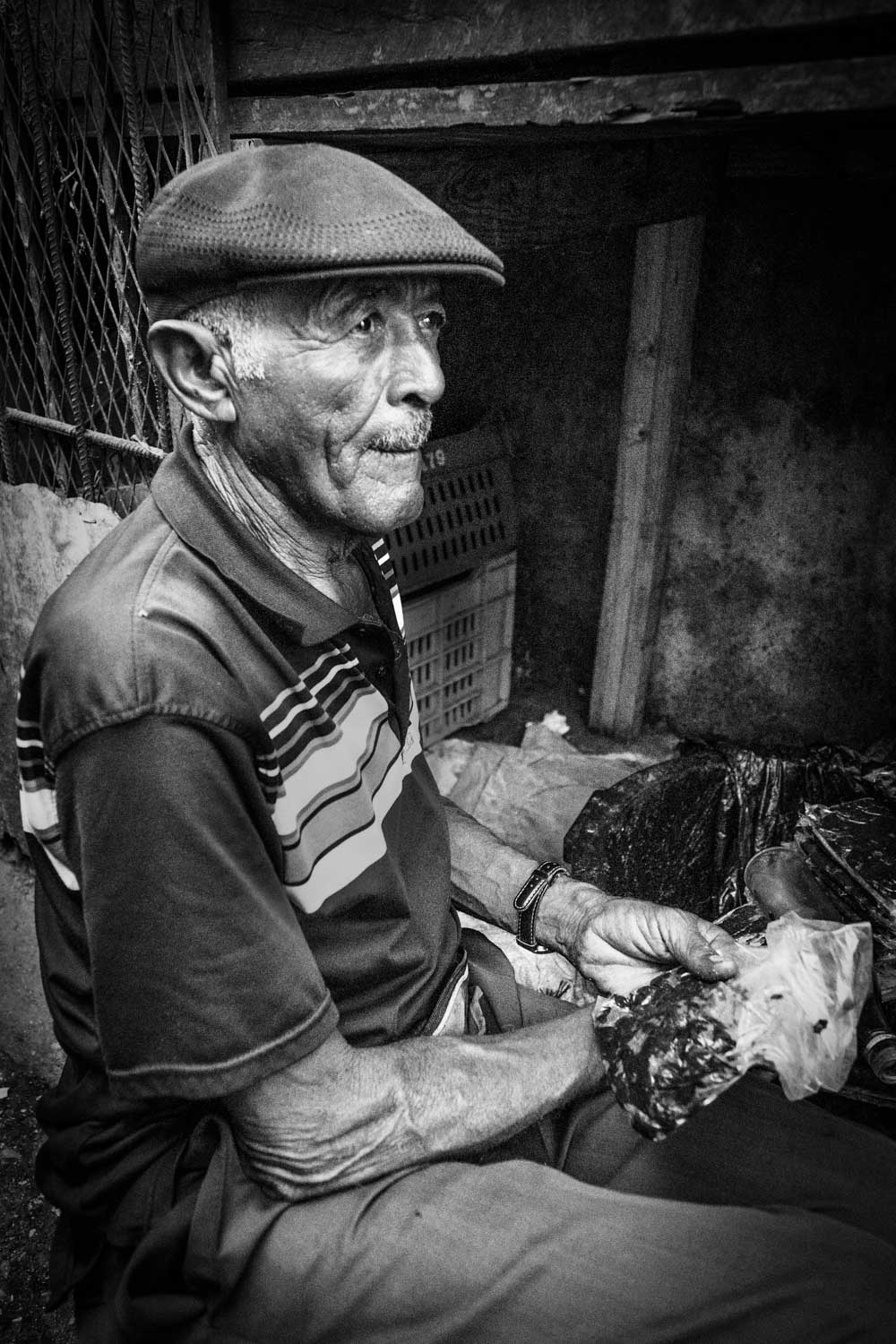 Elderly man in a striped shirt and flat cap sitting thoughtfully, holding a plastic bag, in a black and white urban setting.
