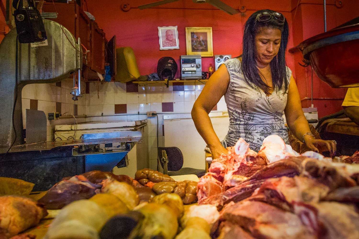 Butcher in a shop cutting various meats on a counter with a red wall background.