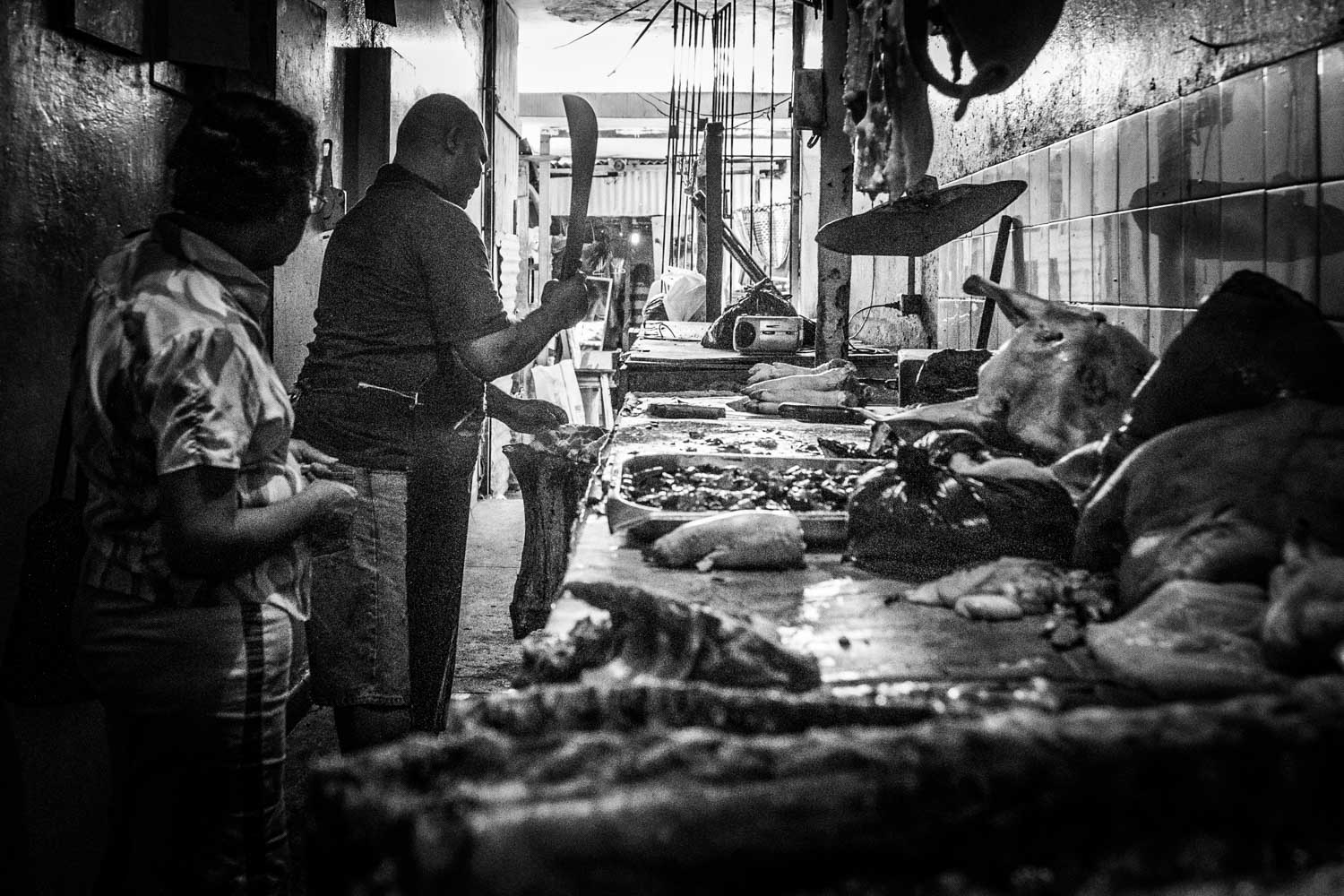 Street butcher shop with meat on display and two people interacting in a narrow, dimly lit corridor.