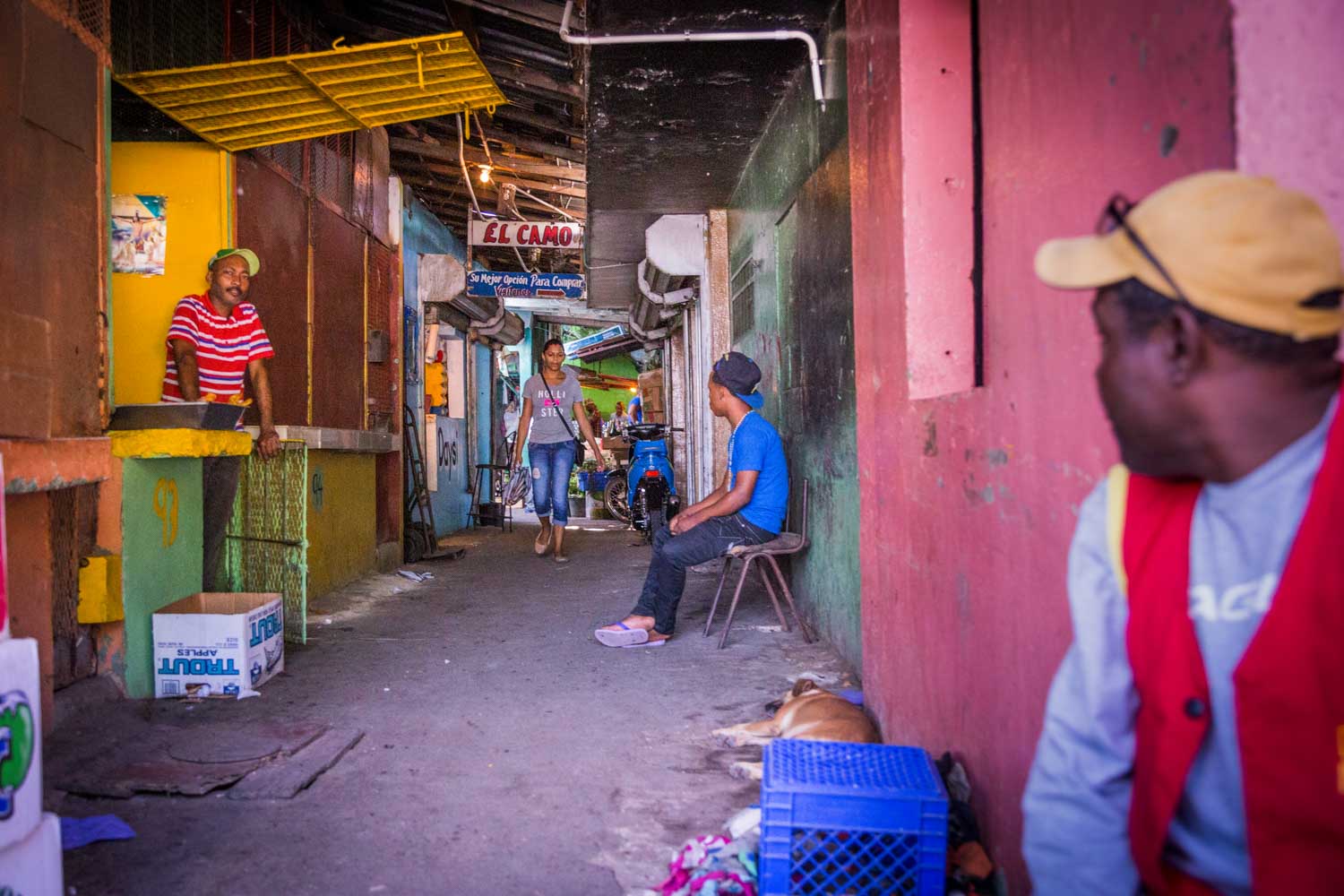 People in a colorful narrow alley with a man in a red shirt, a woman walking, and a man sitting nearby with a dog lying down.