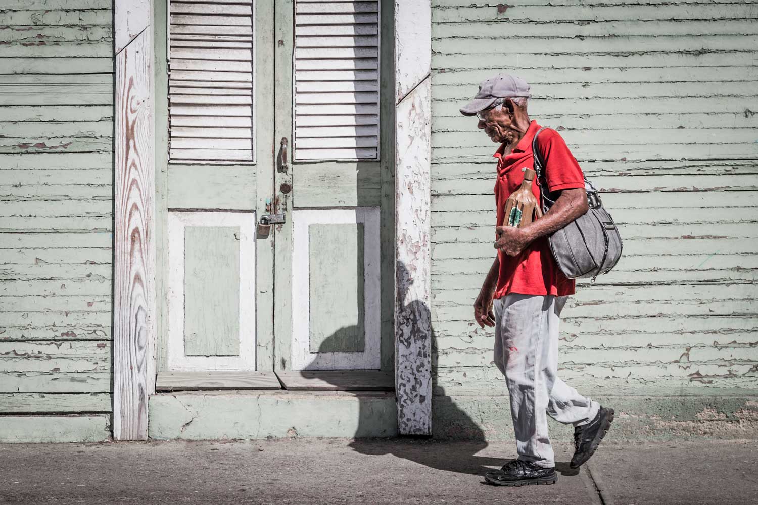 Man in red shirt and cap walking past a weathered green wooden wall, carrying a bag and bottled drink.
