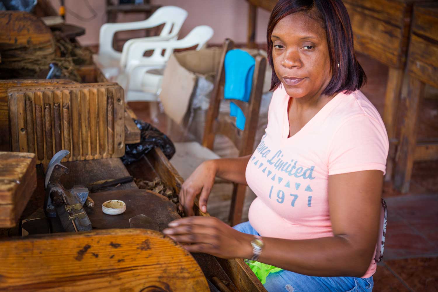 Woman in a pink shirt working with wooden tools in a workshop, seated near white plastic chairs.