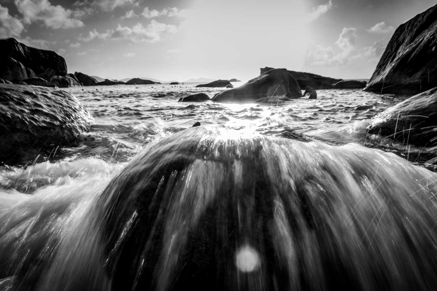 Black and white photo of flowing water over rocks, with sunlight reflecting on the sea and clouds in the sky.