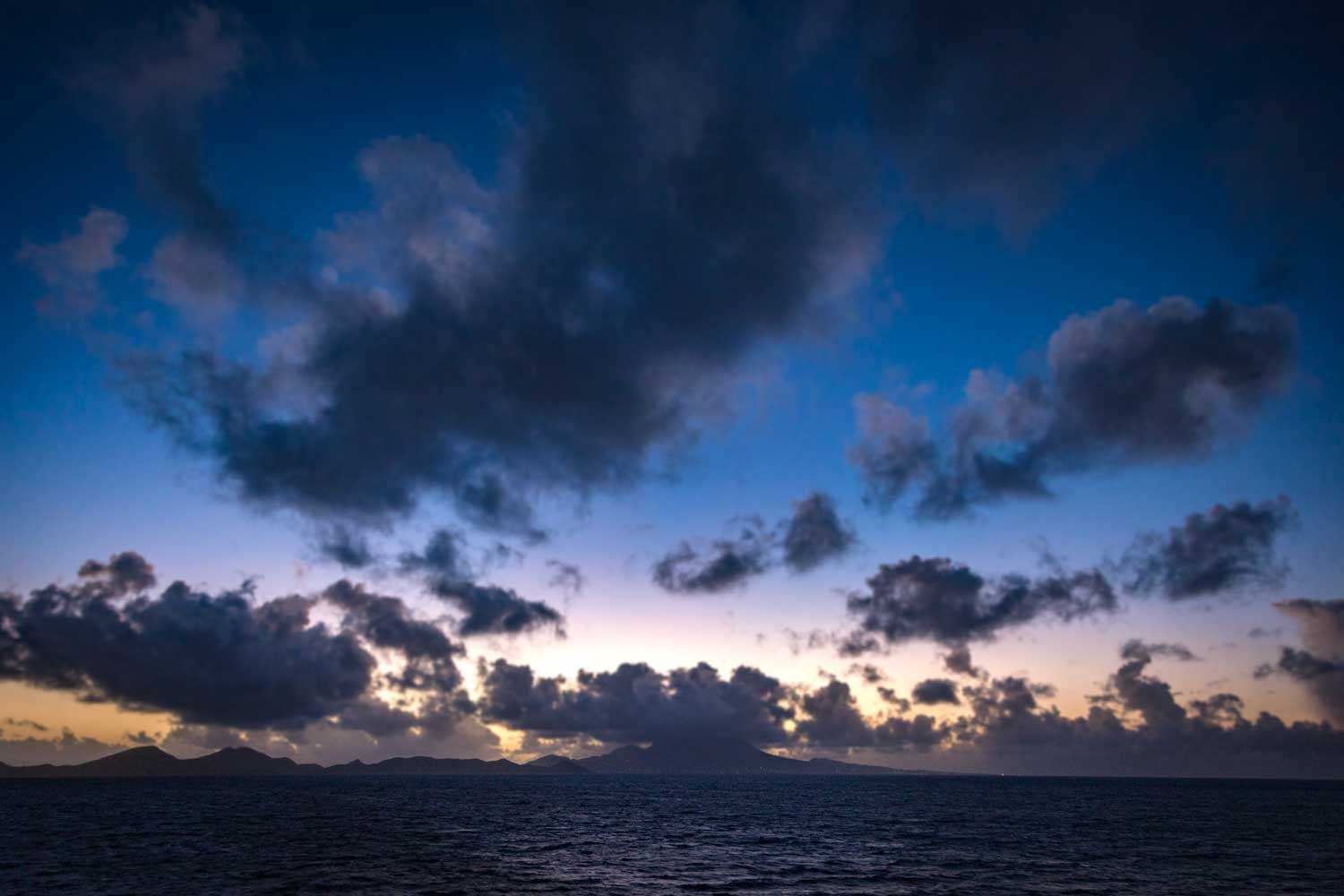 Dramatic sunset over dark ocean with silhouetted islands and scattered clouds in blue and purple sky.