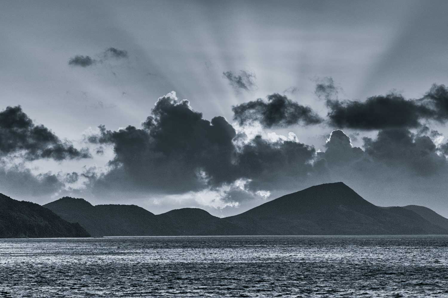 Sun rays break through clouds over silhouetted mountains and a shimmering ocean, creating a dramatic monochrome seascape.