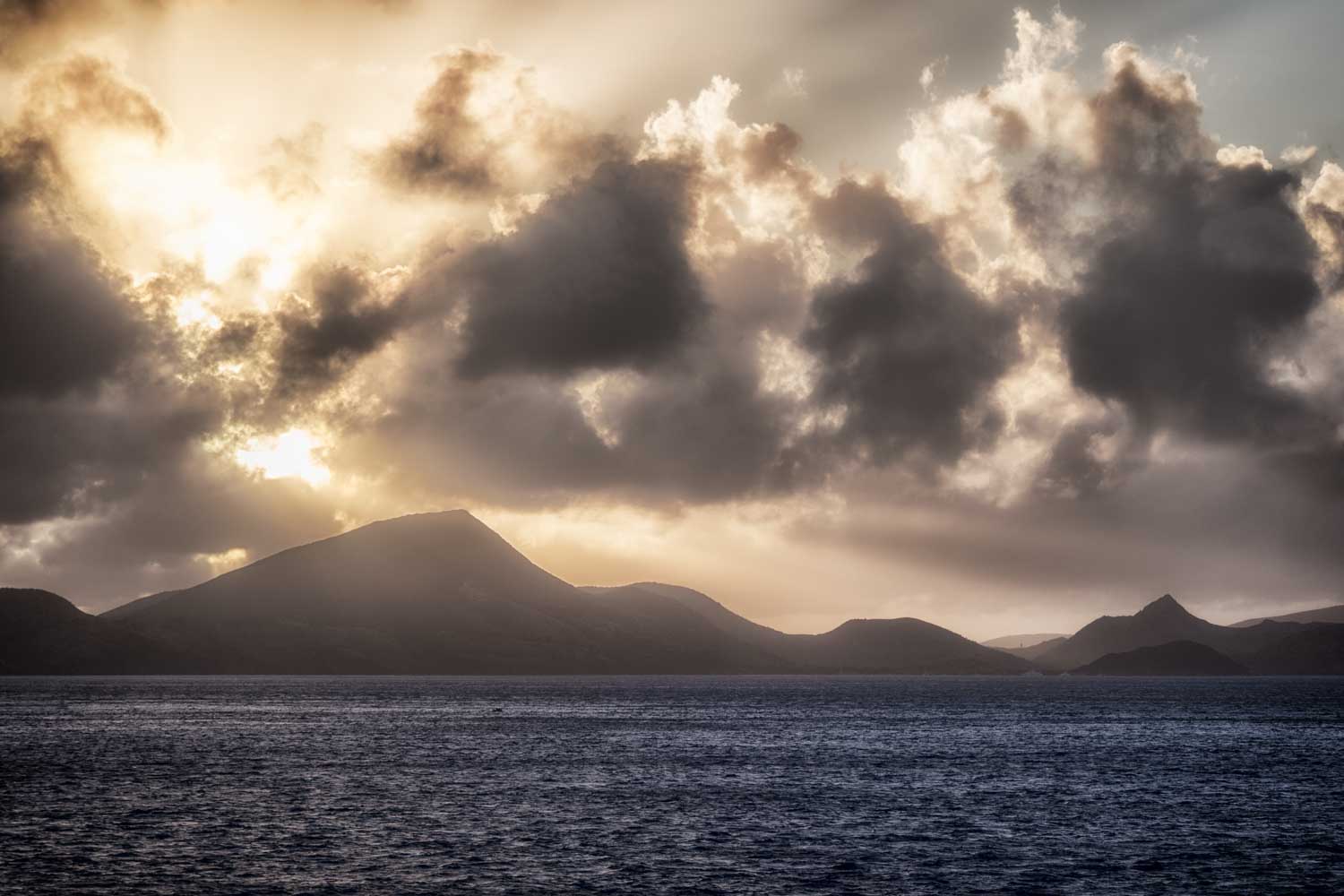 Dramatic sunrise over mountains and ocean, with clouds and sunbeams creating a serene, atmospheric seascape.
