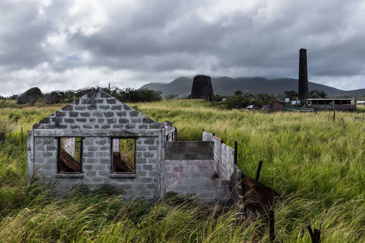 Old stone building ruins surrounded by tall grass, with cloudy mountains and industrial structures in the background.
