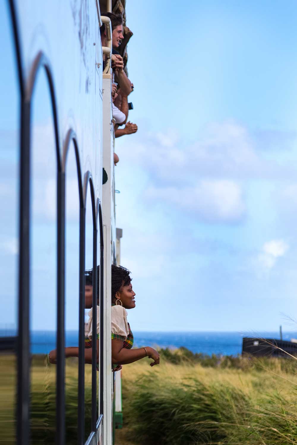 People leaning out of a train, enjoying a scenic view of grassy fields and the ocean under a clear blue sky.