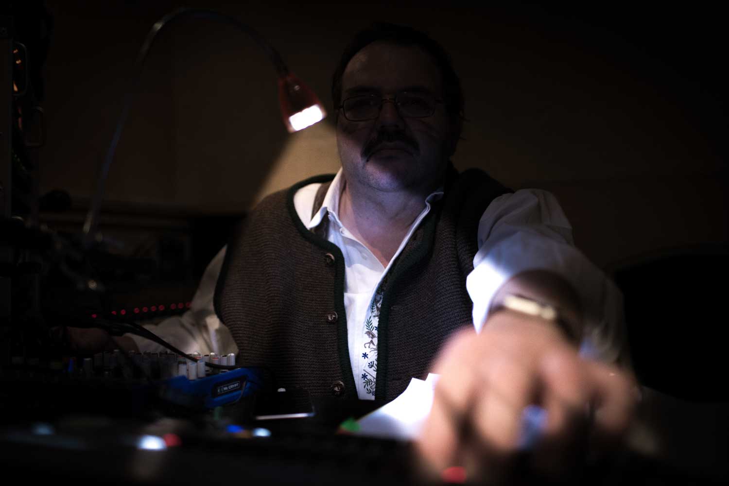 Man in dimly lit room adjusting sound equipment under a small lamp.