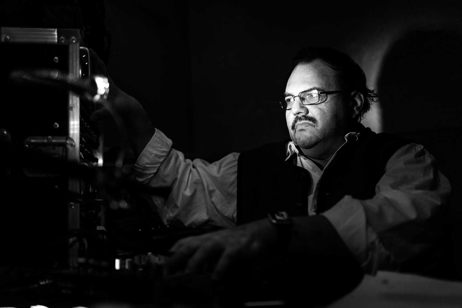 Man in glasses operating audio equipment in dimly lit studio.