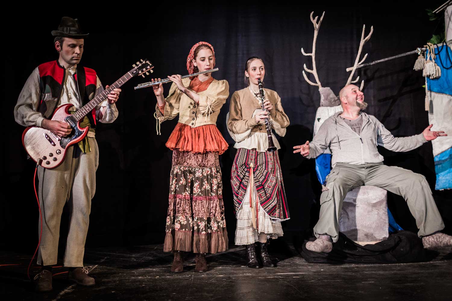 Four musicians in traditional attire perform with guitar, flute, and clarinet on stage featuring a deer backdrop.