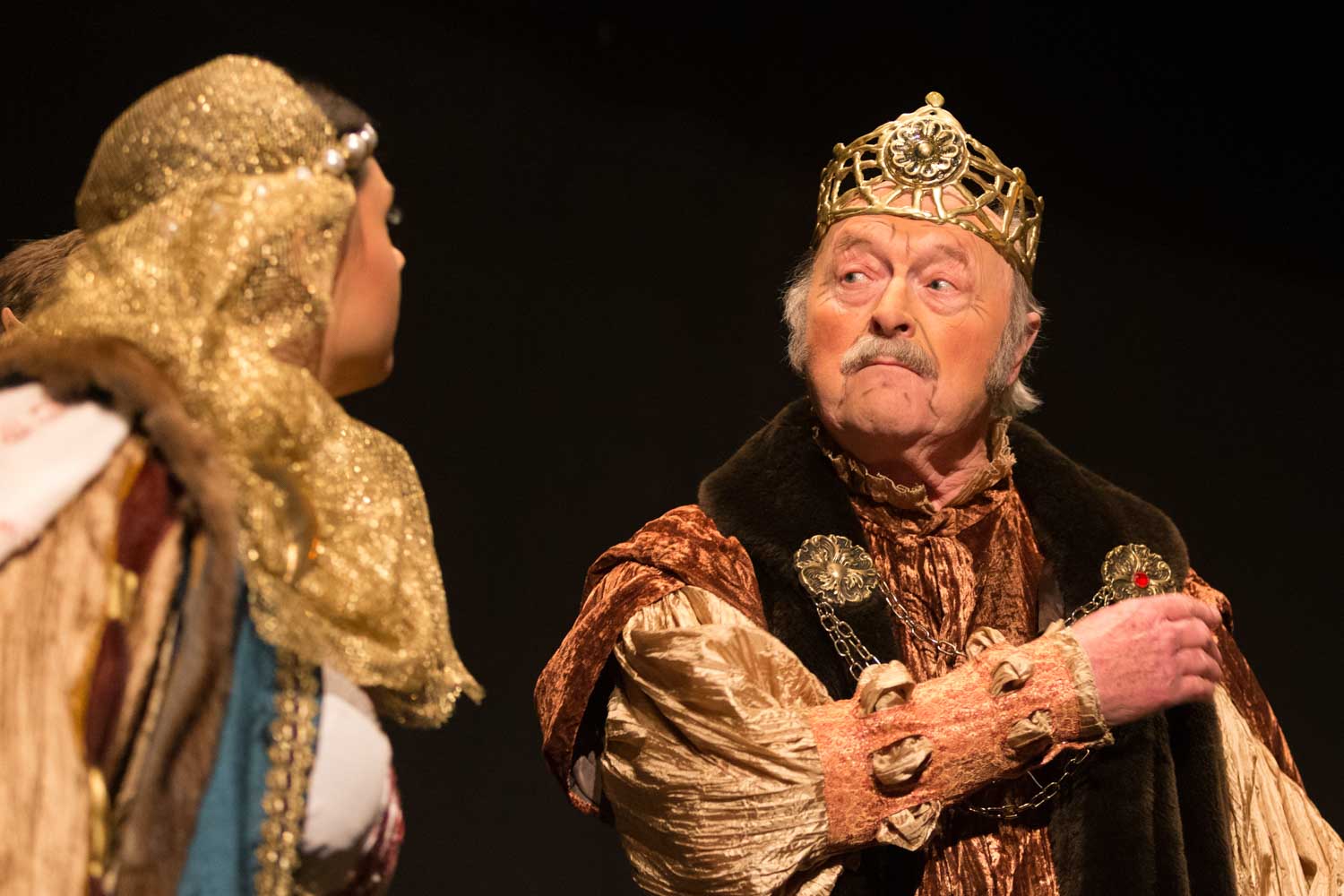 Elderly man in medieval costume with crown on stage, looking at woman in ornate gold headpiece.