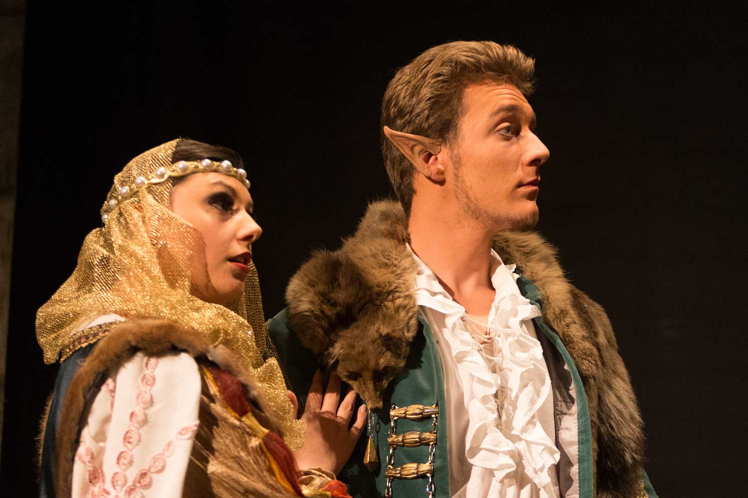 Two actors in medieval costumes performing on stage; one with elf ears and a fur-trimmed coat, the other in a gold headdress.