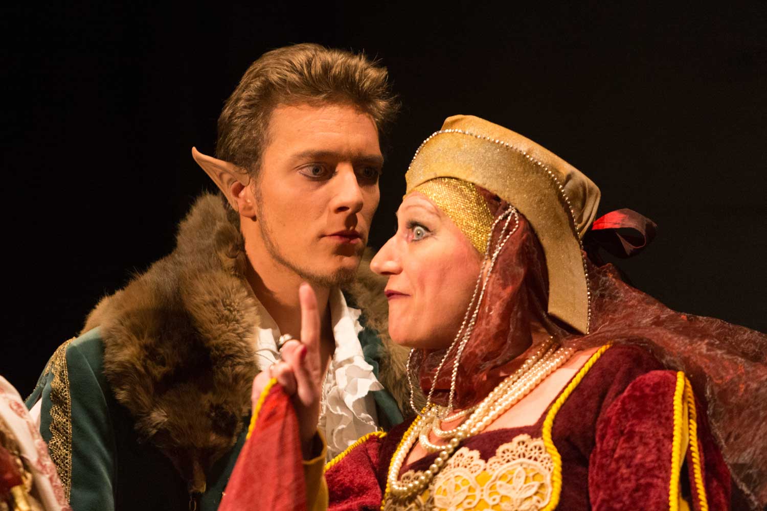 Two actors in medieval costumes perform a dramatic scene, one with elf ears, under theatrical lighting.
