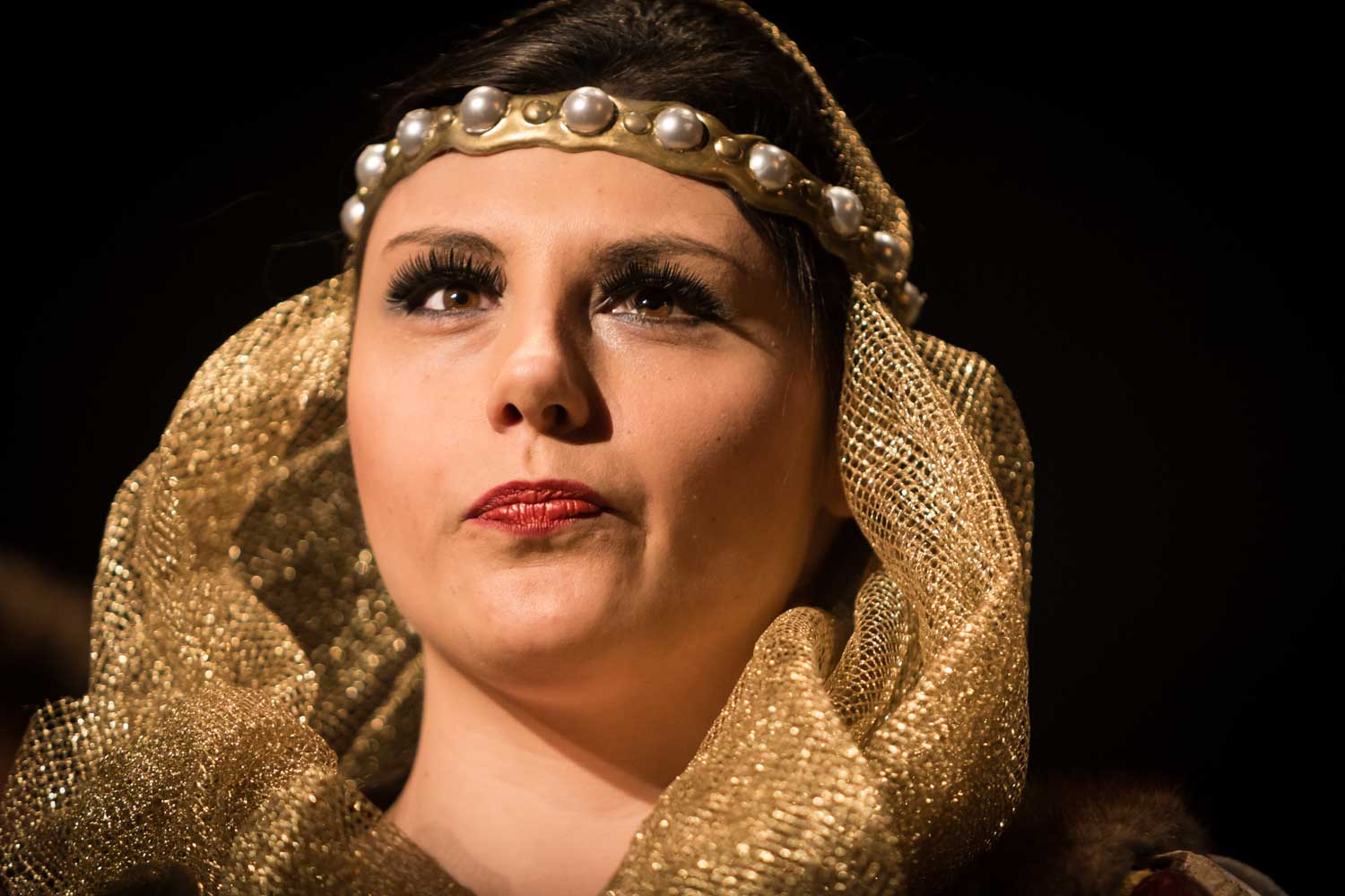 Woman in Renaissance costume with gold headdress and pearl headband, gazing intently in dramatic lighting.