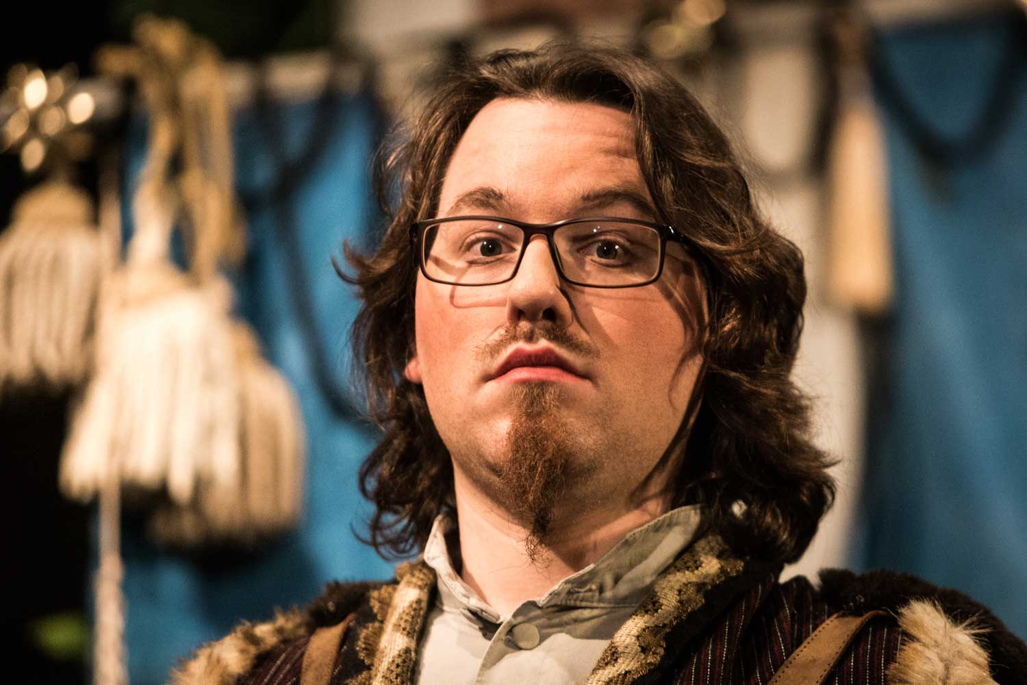 Man with long hair and glasses wearing historical costume in front of blue backdrop.