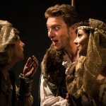 Three actors in medieval costumes perform expressively in a theatrical scene with dim lighting.